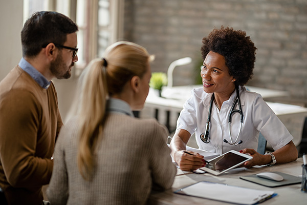 A caregiver, wearing a stethoscope, displays what is on her tablet to the couple sitting on the other side of her desk.