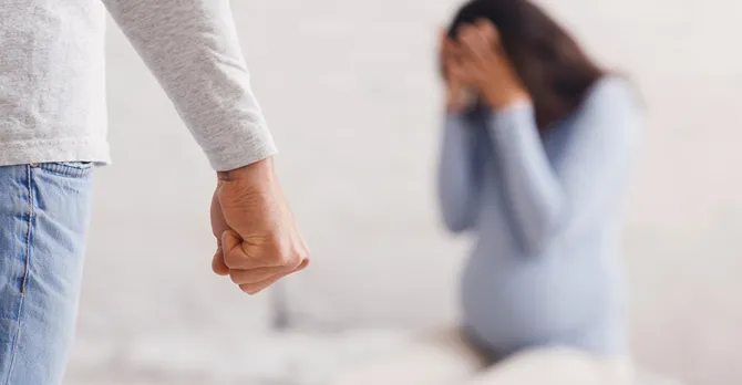 A man's clenched fist is across the room from a scared pregnant woman sitting on the foot of a bed.