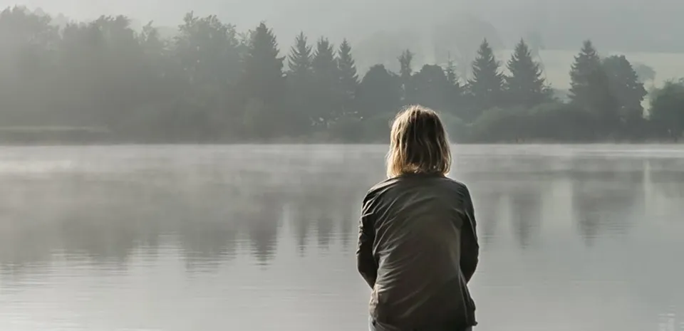 A woman sits on a dock, looking across the water through a thick fog.