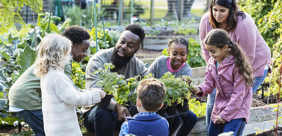 A black man teaches a diverse group of young children about gardening in a community setting.