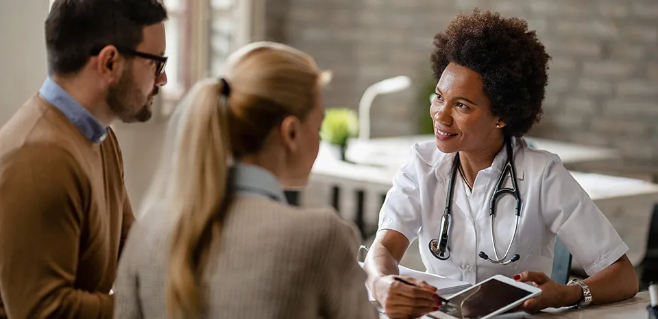 A caregiver, wearing a stethoscope, displays what is on her tablet to the couple sitting on the other side of her desk.