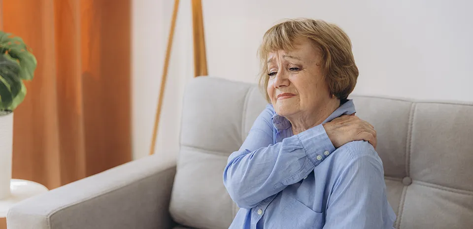 Woman sitting on a couch holding left shoulder wondering if her pain could mean a heart attack