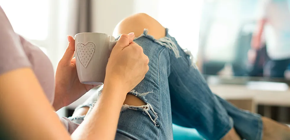 A woman sits in a lounging position as she holds a hot beverage in a cup with a heart (view from behind and to the right)