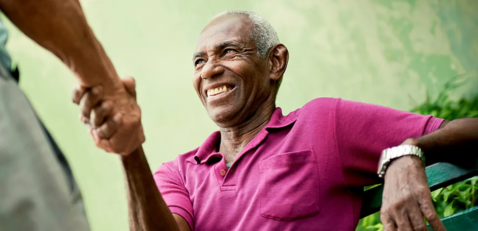 Senior African American man, sitting on bench in park, shakes hands with someone in front of him. 