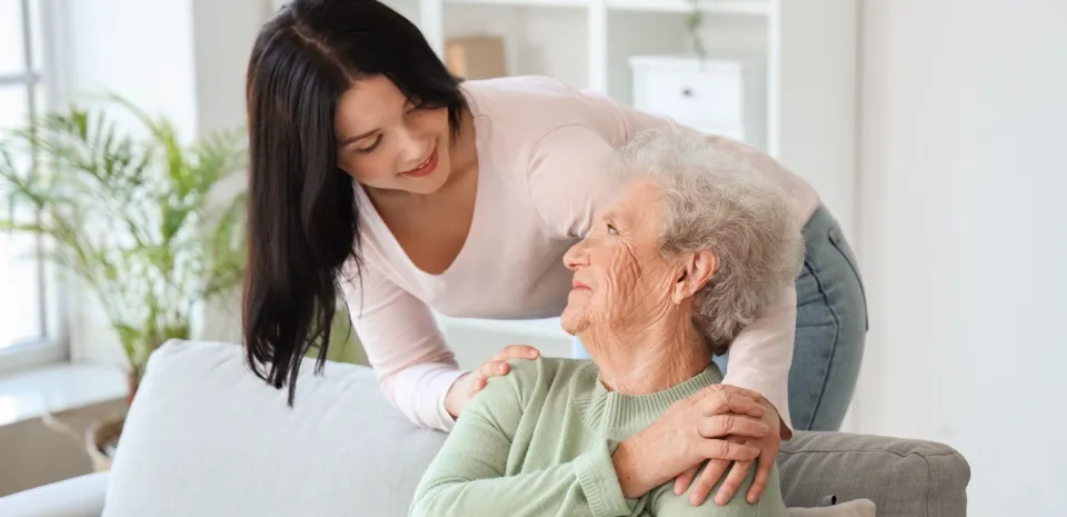 daughter standing behind a couch hugging grandmother while ensuring she is in a safe home