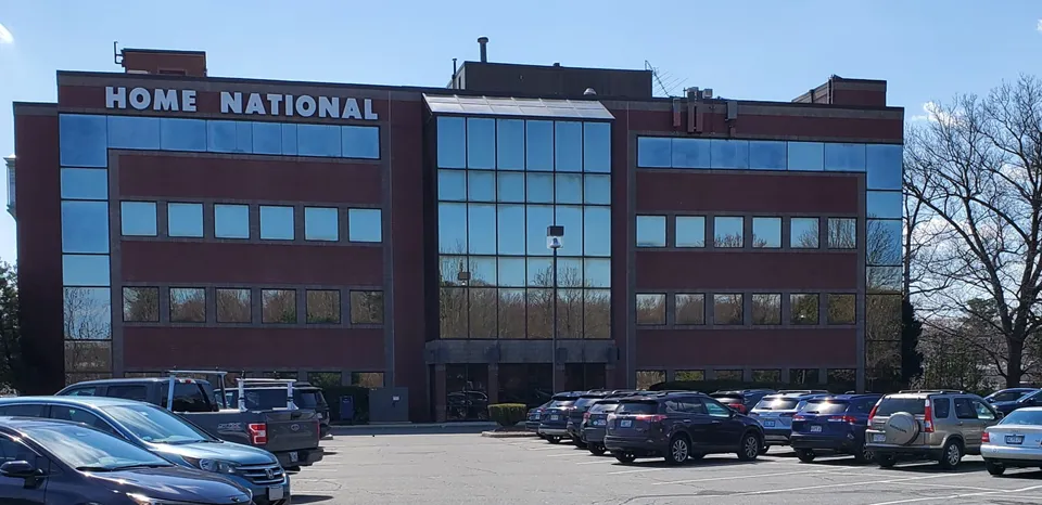 The exterior of a brick building. Many cars are in the parking lot. A sign outside the building reads "Home National."