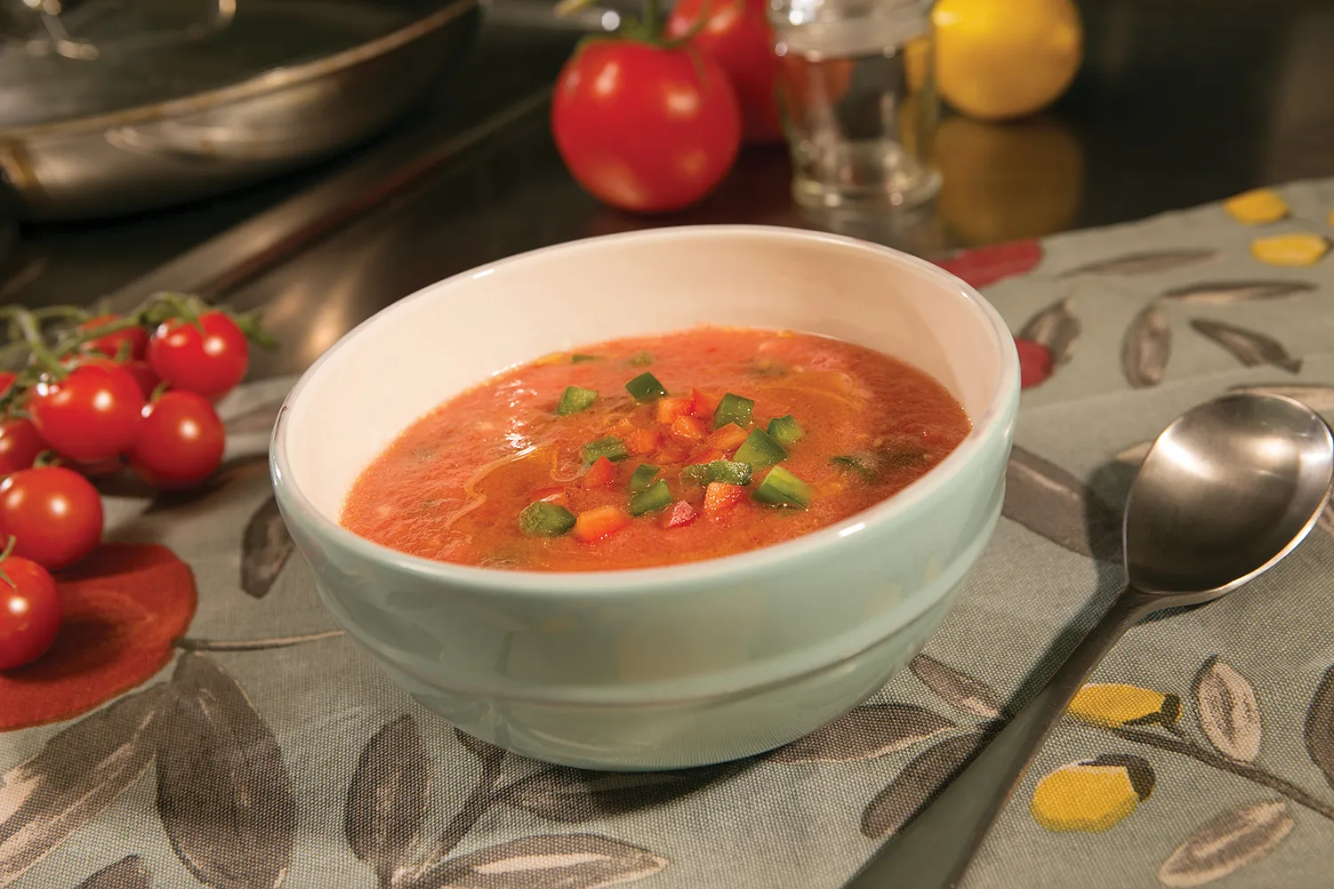 A bowl of gazpacho is is sitting next to a spoon and cherry tomatoes.