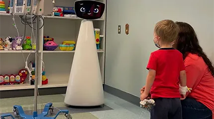 A pediatric patient with an IV drip stands, facing a smiling Robin the Robot, pictured in front of a toy shelf.