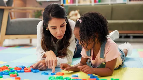Child Life specialist laying on the floor pointing to alphabet letters with a young black child right next to her