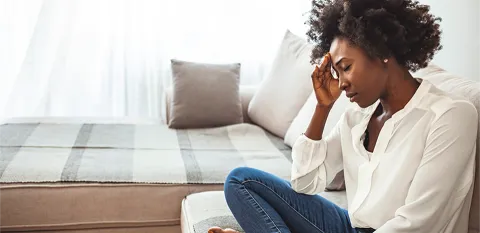 I black woman sitting cross-legged in a chair, hand on forehead and eyes closed.