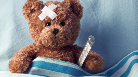 A stuffed teddy bear is in a hospital blanket with a thermometer and bandage on its head.