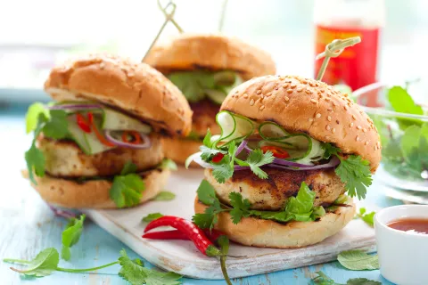 Several turkey burgers with various vegetables on sesame seed buns are shown on a cutting board.