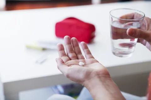 A closeup of a hand holding a white pill in front of a glass of water and a white table.