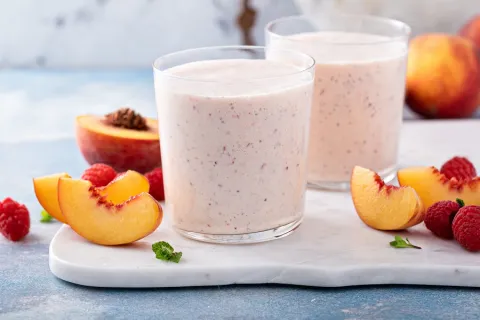 Two smoothie glasses are surrounded by peaches and other berries.