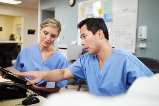 Two nurses in blue scrubs are looking at a computer in a hospital.
