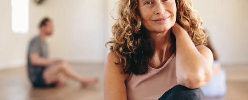 A woman with long hair smiles while sitting on the floor of a yoga, one knee to her chest, one hand behind her neck.