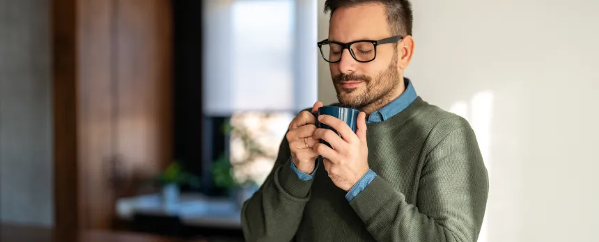 Man with eyes closed holding a warm cup of coffee in a kitchen practicing mindfulness