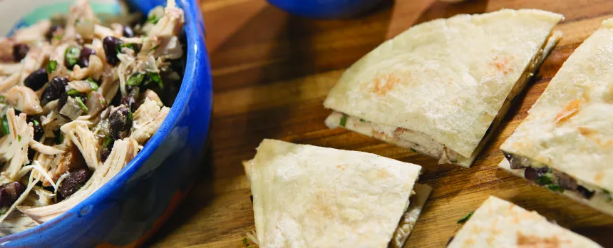 A blue bowl of shredded chicken and black beans is next to a quesadilla that is cut into quarters.