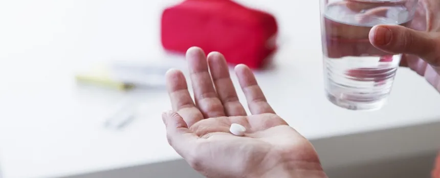 A closeup of a hand holding a white pill in front of a glass of water and a white table.
