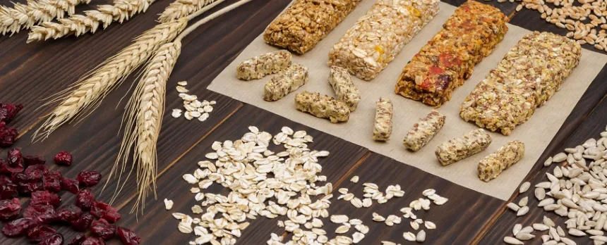 Multiple granola bars are on a table with various grains, oats, and wheat.