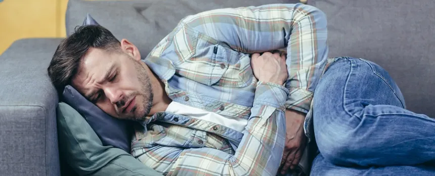 A man lays on a sofa, in the fetal position, while clutching his abdomen.