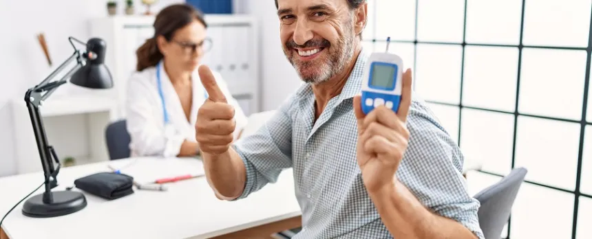 A smiling man is holding a blood glucose meter and giving a thumbs up while in a doctor's office.